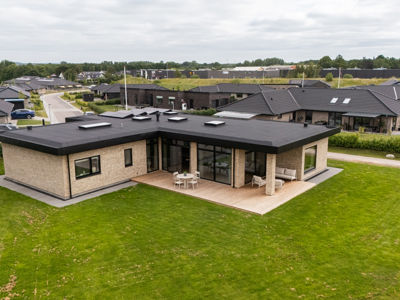 Moderne funkishus fra eurodan-huse med lys murstensfacade, sort fladt tag, store vinduespartier, græsplæne og træterrasse rundt om boligen – inspiration til nybyggeri, husbyggeri og arkitekttegnede husdrømme.